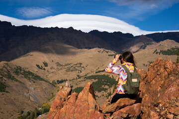 Young Latina woman with sportswear and backpack on top of a hill looking through binoculars at a...