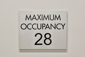 Maximum Occupancy sign on white interior wall in commercial building, 28 capacity copy space. NFPA Fire Marshal requirement.
