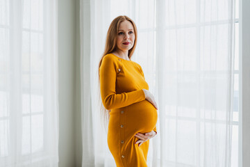 Pregnancy motherhood people expectation future. Pregnant woman with big belly standing near window at home. Girl hugging her tummy enjoying pregnancy. Maternity tenderness parenthood new life concept