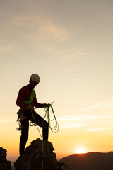 A man in a red jacket is standing on a rock with a green rope. The sun is setting in the...