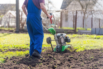 A farmer in the garden tills the land with a motorized cultivator or power tiller, preparing the soil for planting crops