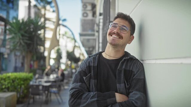 Smiling young man with glasses and moustache leaning against a wall on a sunny urban street.