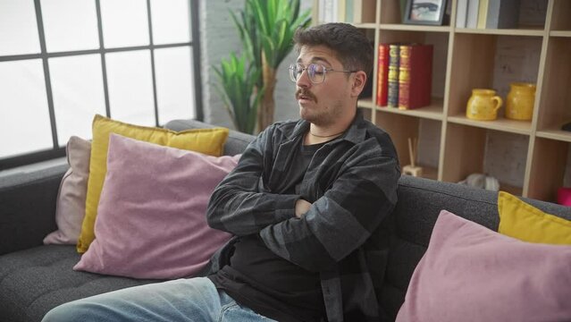 Grumpy young hispanic man with a moustache, skeptically crossed arms on living room sofa, sporting glasses. nervous, disapproving face expression radiates negativity at home.
