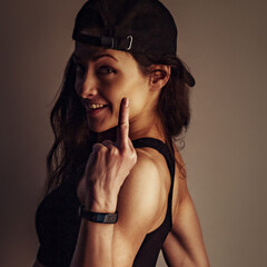 Fuck you. Woman in black sport bra and summer black cap showing the fuck sign the hand, standing back on dark shadow studio background. Closeup