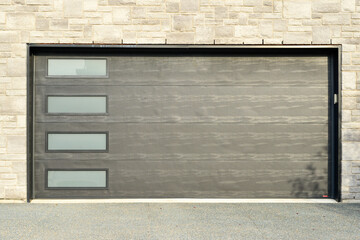 Dark grey double garage door on a residence with tan and beige textured brick. The modern style metal car garage door has four glass panel windows. The exterior automatic door rolls upward.