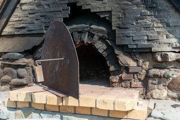 A vintage outdoor wood fired bread oven. The exterior structure is made from brick and rocks with a circular heavy metal door, iron, and wood handle. There's black soot on the hearth of the stove