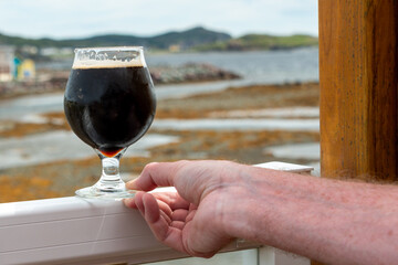 A man holding a stout beer in a clear tulip shaped glass on the railing of a patio. There's a river and grassy marsh in the background. The cold beverage is dark brown with froth on the top.