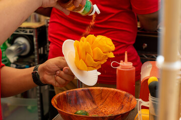 Mexican mango flowers or mango on a stick are made by a street vendor at a market. The fruit is orange color and the vendor is sprinkling lime, chili, salt, and cinnamon over the sticky sweet fruit. 