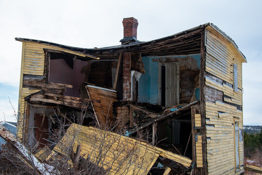 An old yellow wooden house with the exterior wall damaged and demolished. The dilapidated abandoned house is open and exposes the inside rooms of the uninhabited property. The rubble is spread around.
