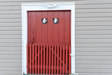 Red exterior wooden panel barn doors with white trim on an old grey building. There are two small horse head signs on the boards of the storage shed. A half gate on hinges is attached to the door. 
