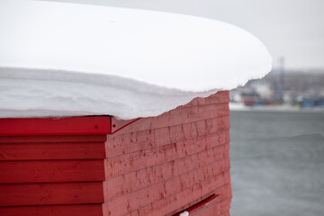 A vibrant red wooden house with a thick heavy drift of fresh white snow. A winter scene of rooftop snow covered with the ocean in the background. The exterior wall is horizontal clapboard siding.