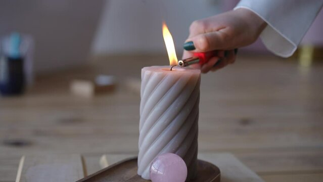 Close-up. A woman's hand with a lighter lights the wick on a large figured wax candle, which stands on a wooden stand with a pink quartz ball indoors