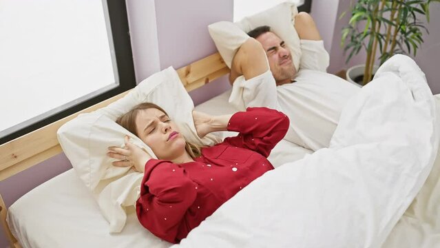 A woman annoyed by snoring lies awake next to a sleeping man in a bedroom setting, depicting a common couple's issue.