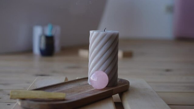 Close-up. A woman extinguishes a burning wick with her fingers on a large figured wax candle, which stands on a wooden stand with a rose quartz ball indoors