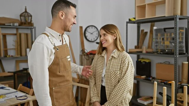 A smiling man and woman in a bright workshop wearing aprons with woodworking tools in the background.