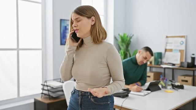 A woman on a phone call in an office with a focused man working at a desk in the background, depicting colleagues in a workplace.