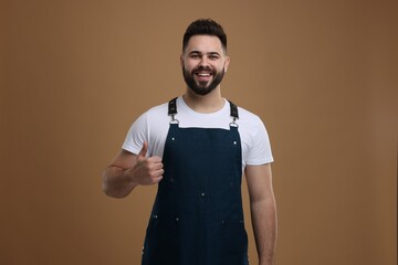 Smiling man in kitchen apron showing thumb up on brown background. Mockup for design