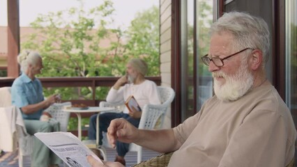 Medium side tilt shot of senior Caucasian male pensioner sitting on terrace in retirement home on summer morning, reading newspaper, pondering global events, taking off glasses and scratching beard - Powered by Adobe
