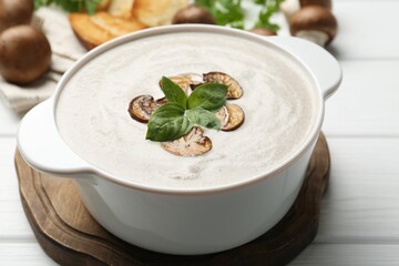 Fresh homemade mushroom soup in ceramic pot on white wooden table, closeup