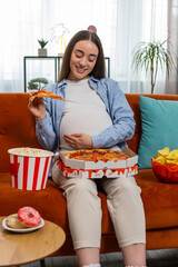 Pregnant woman eating unhealthy junk food pizza, popcorn crisp sitting on sofa watching TV movie in...