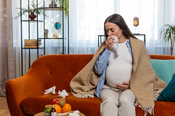 Pregnant unhealthy Caucasian woman girl wrapped in blanket blowing nose in tissue while sits on...