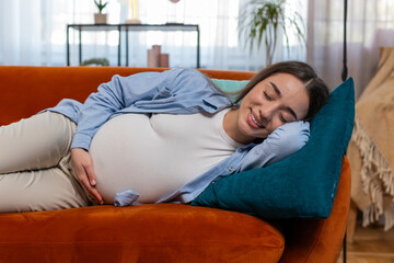 Smiling young pregnant woman stroking big belly while sleeping on comfortable sofa dreaming in...