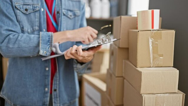 A man in a denim jacket writes on a clipboard in a warehouse with cardboard boxes stacked behind him.