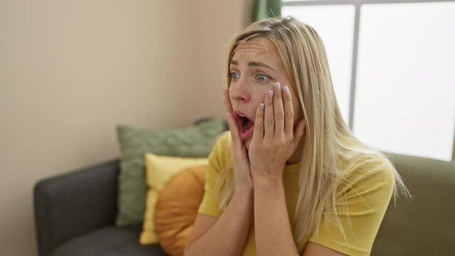 Beautiful young blonde woman at home, wearing a t-shirt, expresses shock and fear through her amazed and surprised face