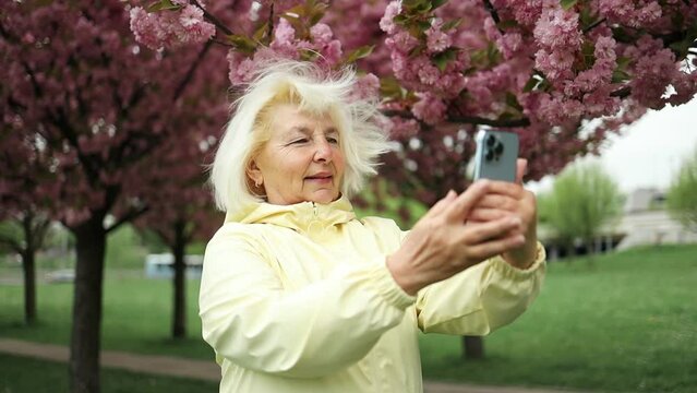 Senior blonde woman traveler makes photos on a smartphone while walking in spring sakura garden and enjoying nature, travel and summer adventures. High quality photo