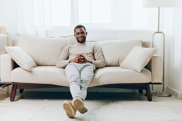 Relaxed African American man sitting on a comfortable sofa in his modern living room, smiling while...