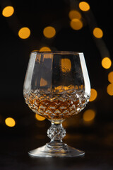 A glass of liquor is sitting on a table. The glass is half full and the liquid inside is brown. The image has a moody and mysterious feel to it, as the light is dim