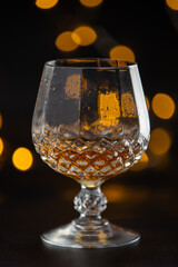 A glass of alcohol is sitting on a table. The glass is half full and has a yellowish tint. The image has a moody and mysterious feel to it, as the focus is on the glass and the liquid inside it