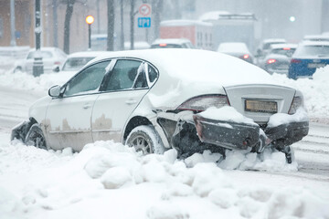 A damaged car covered in snow is stranded on a snowy road during a heavy snowfall, with blurred traffic signs and falling snowflakes visible in the background.