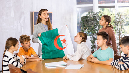 Smiling young woman teacher showing state flag of Algeria and telling preteens schoolchildren...