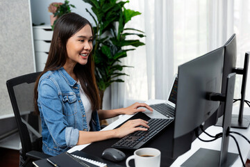 Smiling young Asian IT developer working software development information on pc screens and laptop,...