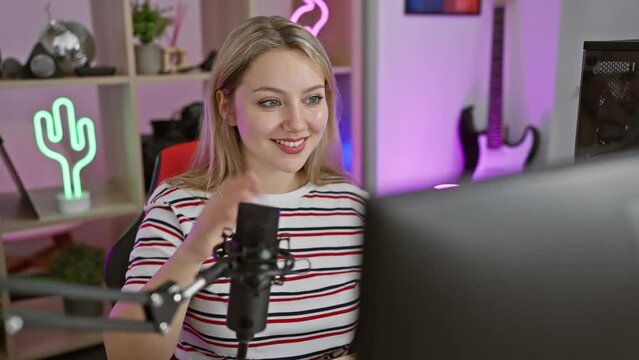 Joyful young blonde streamer saying hello with a friendly gesture, flashing a radiant smile in her gaming room while streaming on computer
