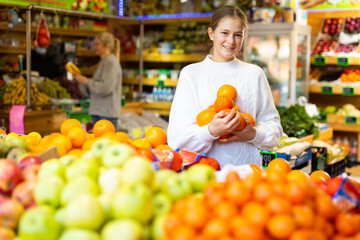 Cheerful teen girl shopping for organic fruits, choosing sweet ripe oranges in local food store ..