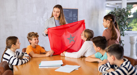 Kindly pedagogue demonstrating Morocco flag to preteen pupils in teaching room during class