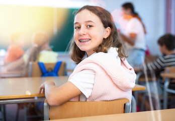 Portrait of smiling schoolgirl with braces sitting at desk in classroom and studying during lesson.