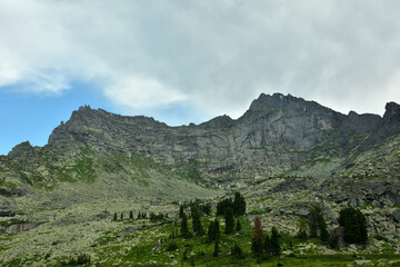 The ridge of a high sheer rock in a mountain range with rare cedars on the slope under a summer cloudy sky.