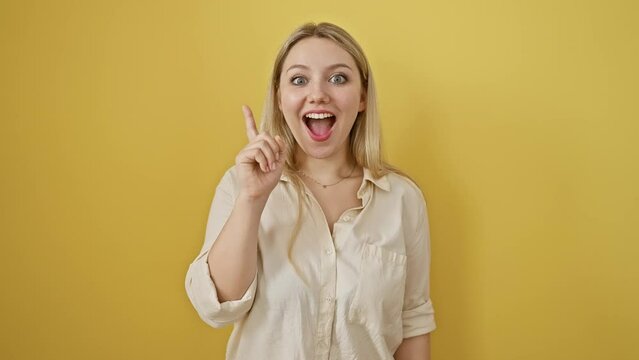 Chuffed to bits, successful young blonde woman stands pointing number one finger up, basking in her clever idea. isolated against a vibrant yellow background, her joy is contagious!