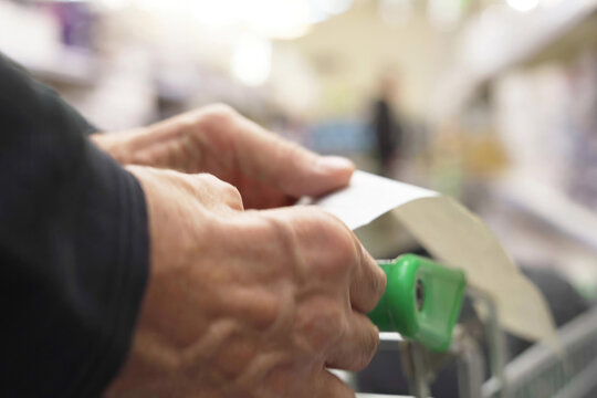 The Hands Of An Unrecognizable Older Man Hold A Sales Receipt Next To A Grocery Cart In A Supermarket.