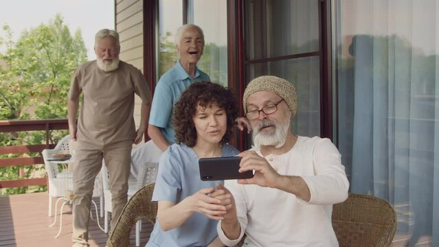 Medium footage of senior Caucasian man and nurse sitting together on terrace, calling for companions to come, posing together, smiling and taking selfie on smartphone, then looking at photo