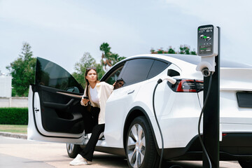 Young woman use smartphone to pay for electricity at public EV car charging station green city park. Modern environmental and sustainable urban lifestyle with EV vehicle. Expedient - obrazy, fototapety, plakaty