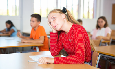 Portrait of tired teen girl sitting with head resting on hand at lesson in school, listening to...
