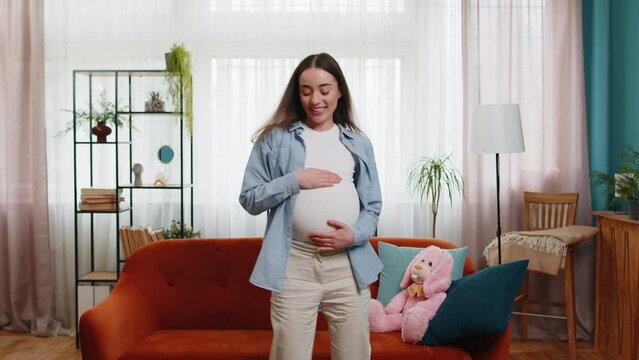 Joyful young pregnant woman full of natural happiness and confidence enjoys playfully dancing and tenderly touching her big belly all while flashing friendly, carefree smile. Future mother at home.