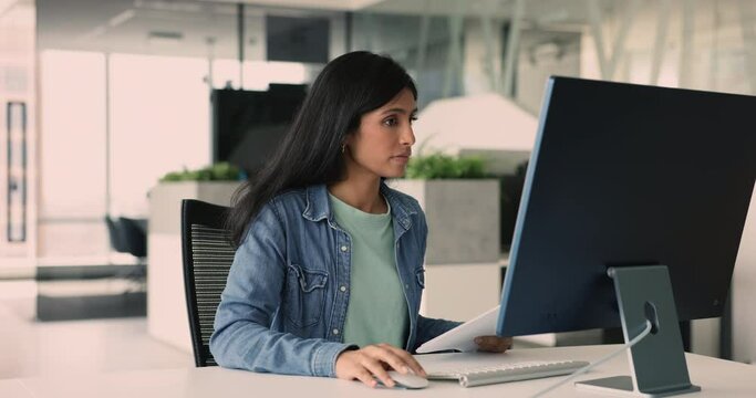 Female employee working in office, sits at desk analyzing data and compiling reports based on information, typing on PC keyboard, drafting or editing documents, prepare report or marketing materials