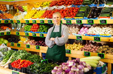Confident successful female grocery store owner in apron standing near shelves of fruits and...