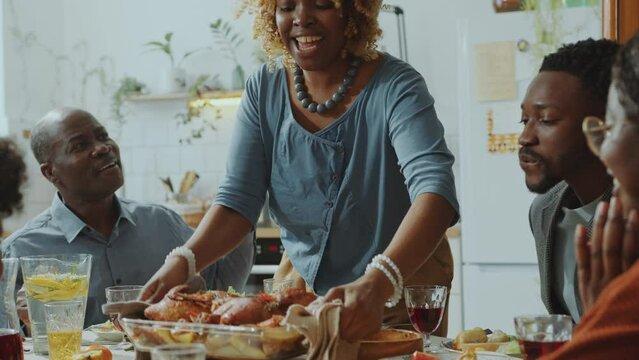 Cheerful African American woman serving dish with baked chicken and potatoes on dinner table, taking seat and speaking with family members on home celebration