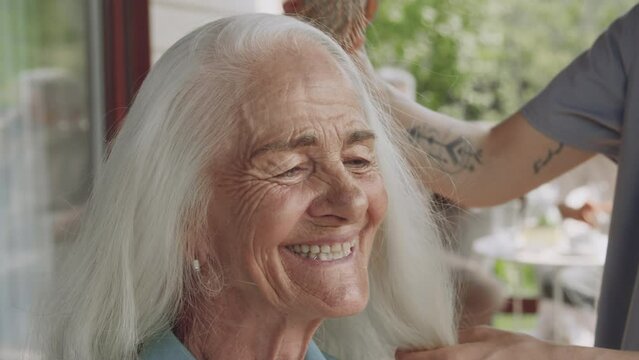 Close-up footage of beautiful 80-year-old Caucasian woman with wrinkly face smiling while chatting to unrecognizable female nurse with tattooed hands, in uniform, who is gently brushing her hair